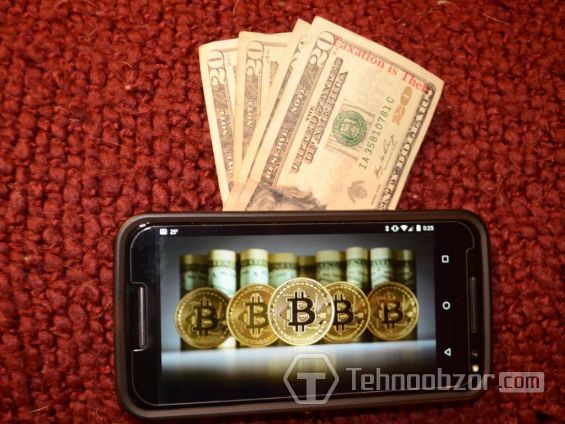 Монети біткоіни на екрані смартфона, що лежить на 20-доларових купюрах